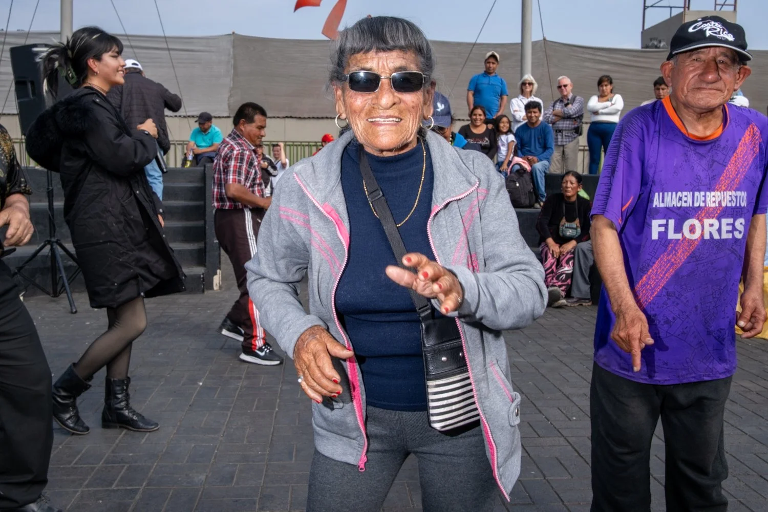 A smiling elderly woman with sunglasses and gray hair, wearing a gray jacket over a navy shirt, standing outdoors among a group of people. She has earrings, a gold necklace, and is holding her hand up in front of her. Behind her, there are other peop