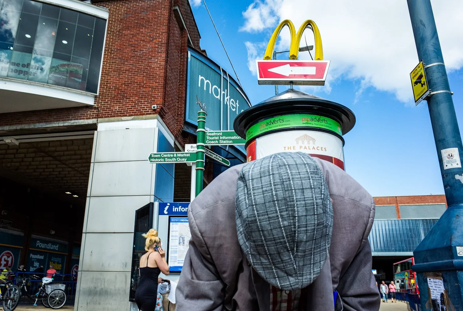 Person wearing a gray plaid hat and gray jacket stands in front of a bus station, with a McDonald's sign and a blue building with signs for the market and South Quay visible behind. A woman with blonde hair and black shirt talks on her phone near bic