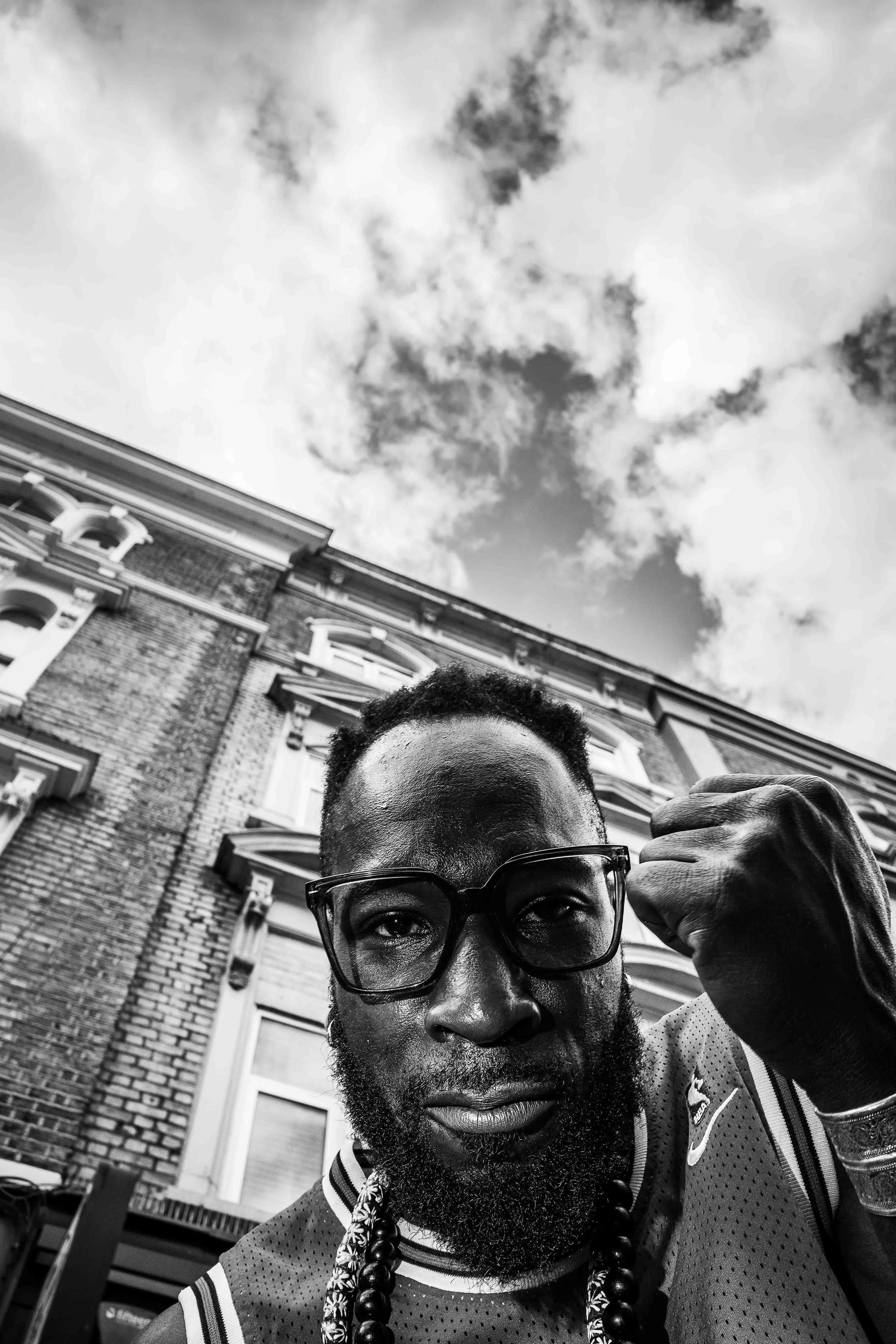 A black and white photo of a man with glasses and a beard, looking at the camera, with a building and cloudy sky in the background.
