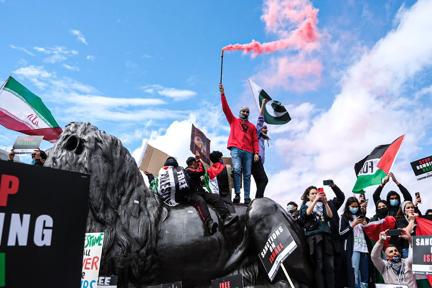 Protesters gather around a large statue of a lion's head during a demonstration. Some individuals are holding national flags, including Iran and Palestine, and signs supporting Palestine. One person is standing on the statue holding a flare that rele