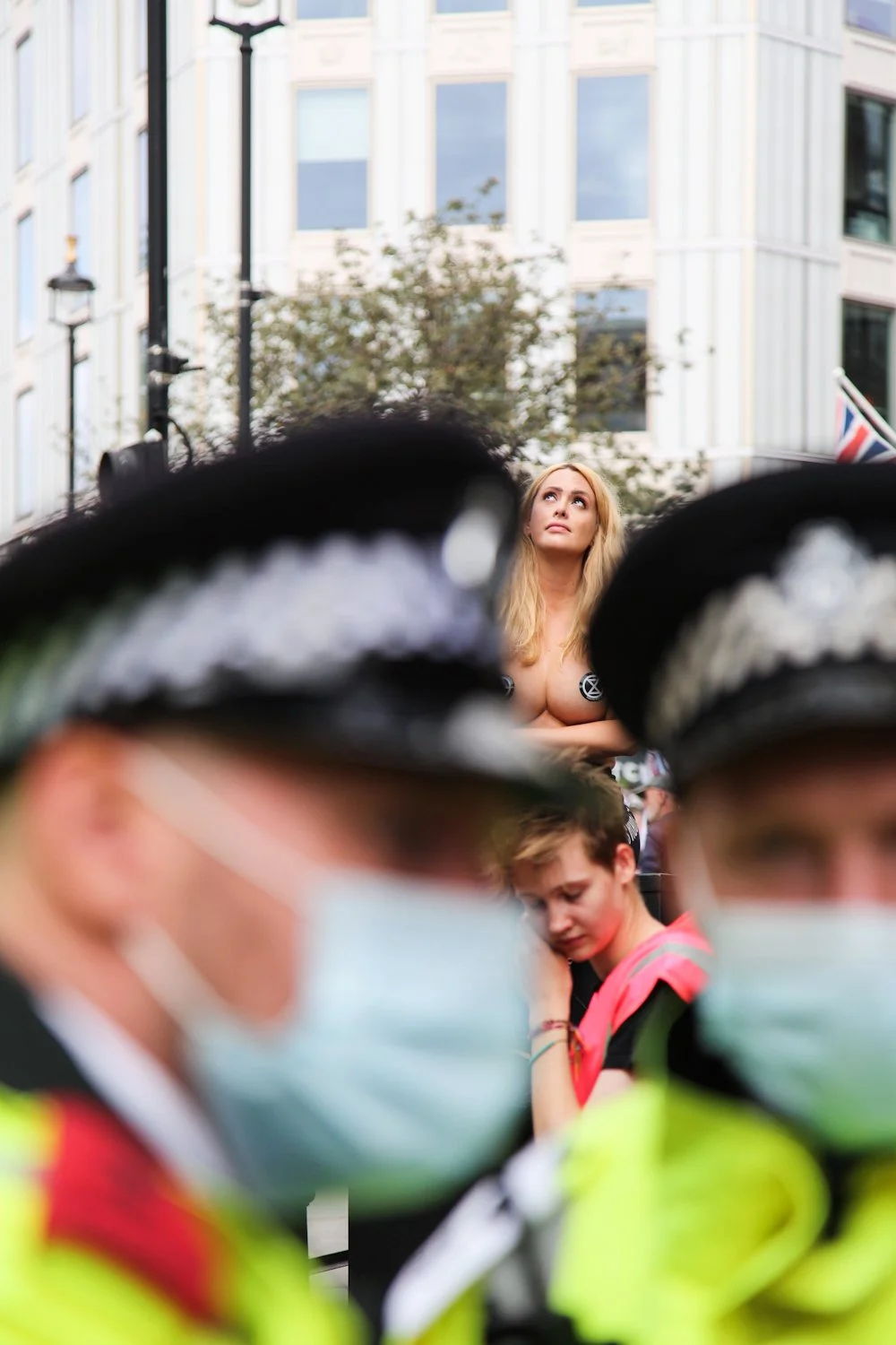 A woman with long blonde hair looking upward, seen between police officers wearing masks and helmets, in an outdoor urban setting.