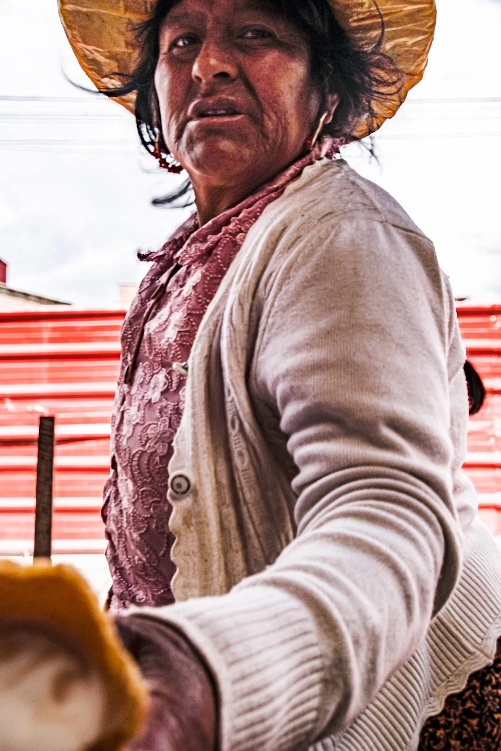 A woman wearing a wide-brimmed straw hat and a light-colored sweater, standing outdoors with a red fence and cloudy sky in the background.