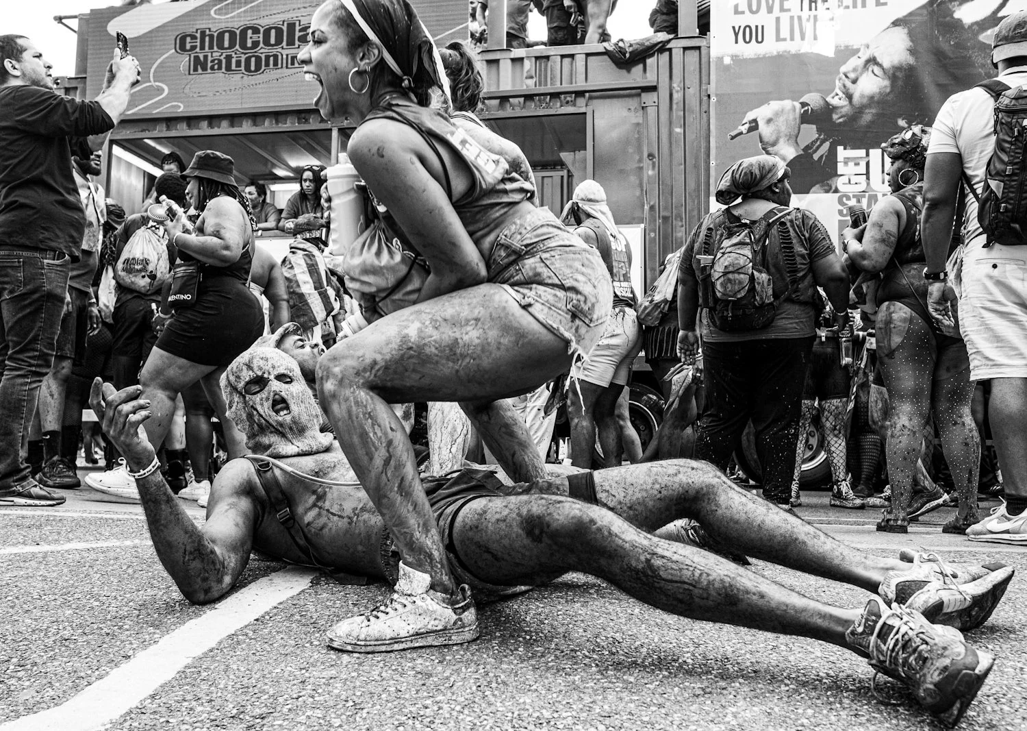 Black and white photo of a street protest with people covered in mud. A person wearing a mask lies on the ground while another person is in a squatting position over them, both covered in mud. The background shows a crowd of people, some taking pictu