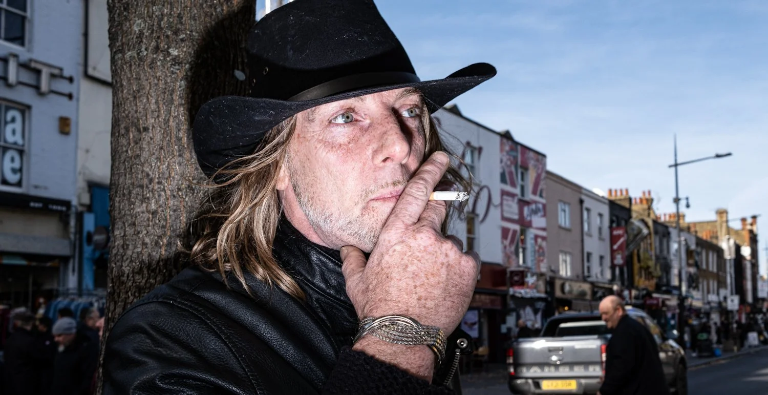 A man with long hair, wearing a black leather jacket, a black cowboy hat, and a silver watch, leaning against a tree, smoking a cigarette on a busy street with shops and pedestrians.