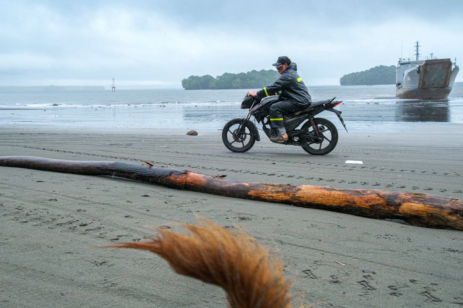 A person wearing a black rain jacket and pants riding a dirt bike on a sandy beach with driftwood, with a large ship in the background and cloudy sky.