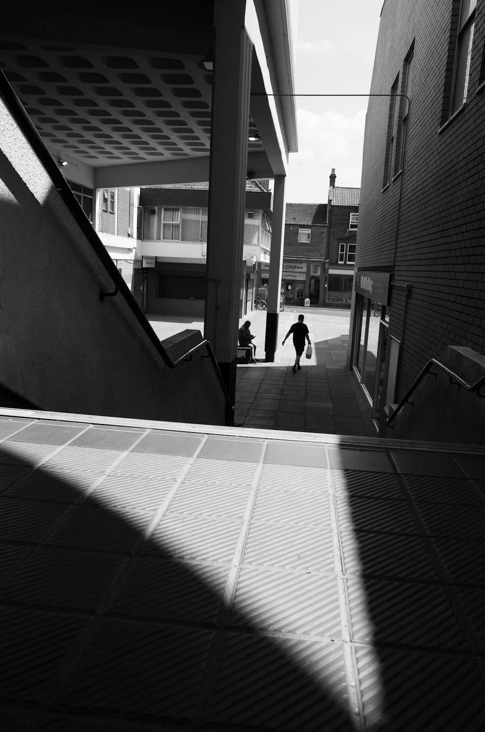 Black and white photo of an urban scene viewed from a staircase, with two people walking outside near shops and buildings.