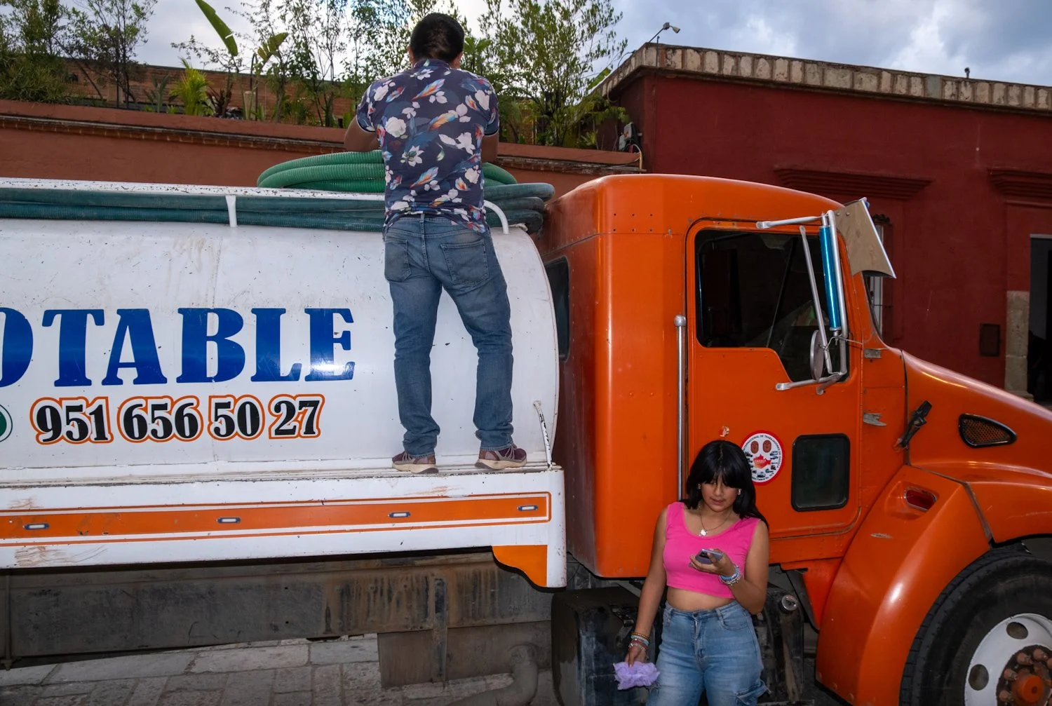 A man standing on the back of an orange and white water delivery truck, wearing a floral shirt and jeans, with a woman in a pink crop top and jeans holding a phone in front of the truck.