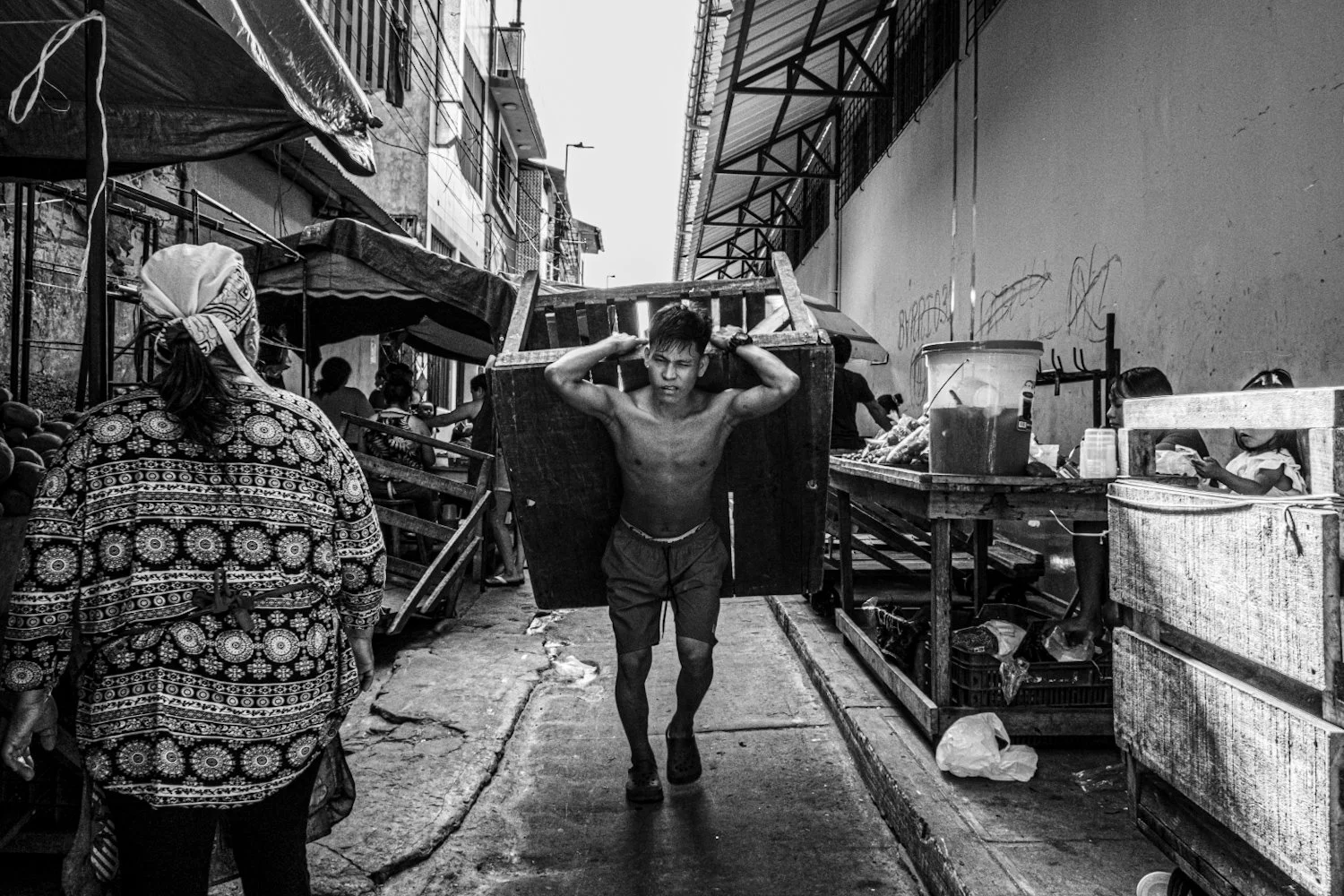 A young man carrying a large wooden crate on his shoulders in a busy marketplace. There are vendors and shoppers, some behind stalls, with buildings and signs visible in the background.