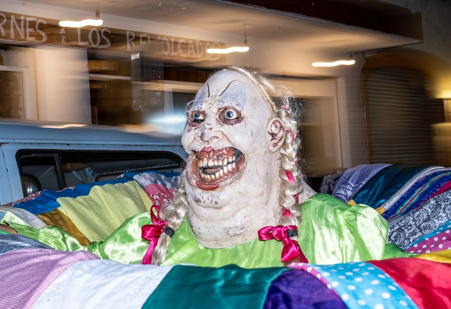 A person wearing a creepy clown mask with exaggerated facial features, blonde braided hair with pink ribbons, and a bright green outfit, surrounded by colorful patchwork blankets in a room with blurred background.