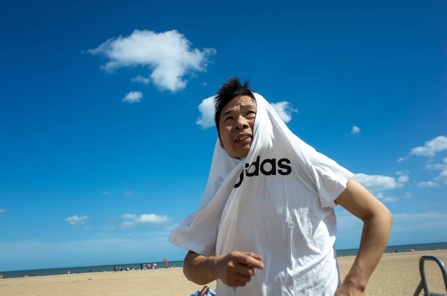 A man standing on a sandy beach with the ocean and blue sky with clouds in the background, wearing a white Adidas t-shirt, holding a white cloth over his head.