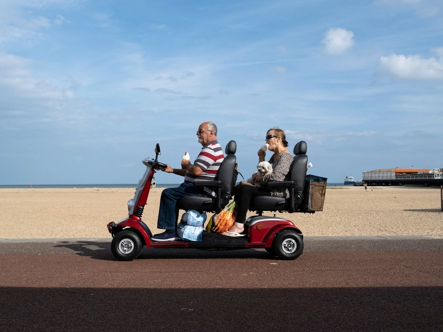 Older couple riding a red electric scooter along the beach road, eating ice cream, with a woman holding a small dog, beach and ships in the background.