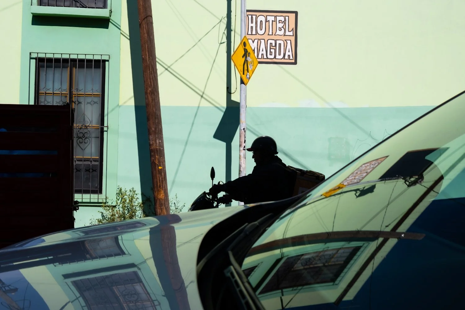 A silhouette of a person riding a motorcycle at street level, with cars in the foreground and a light green building in the background. There is a sign that reads 'Hotel Magda' and a yellow pedestrian crossing sign on a utility pole.