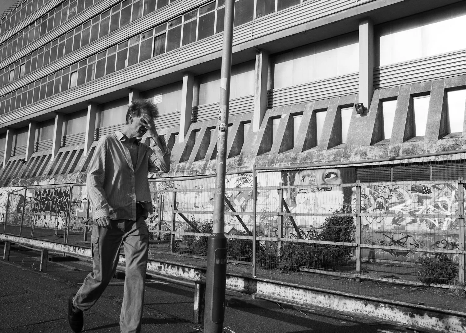 A person walking on a sidewalk in front of a graffiti-covered fence with a large building in the background.
