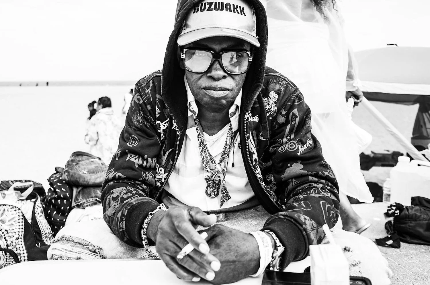 A man wearing large glasses, a cap, and a hooded jacket with graffiti-style patterns sitting on the ground at the beach, with bags and people in the background.