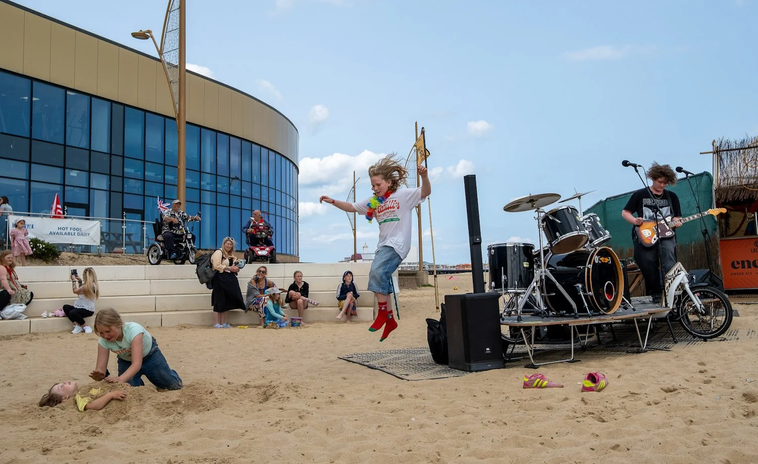 Children playing in sand with a woman jumping, a band playing music nearby, spectators sitting on steps, and a modern glass building in the background on a cloudy day.