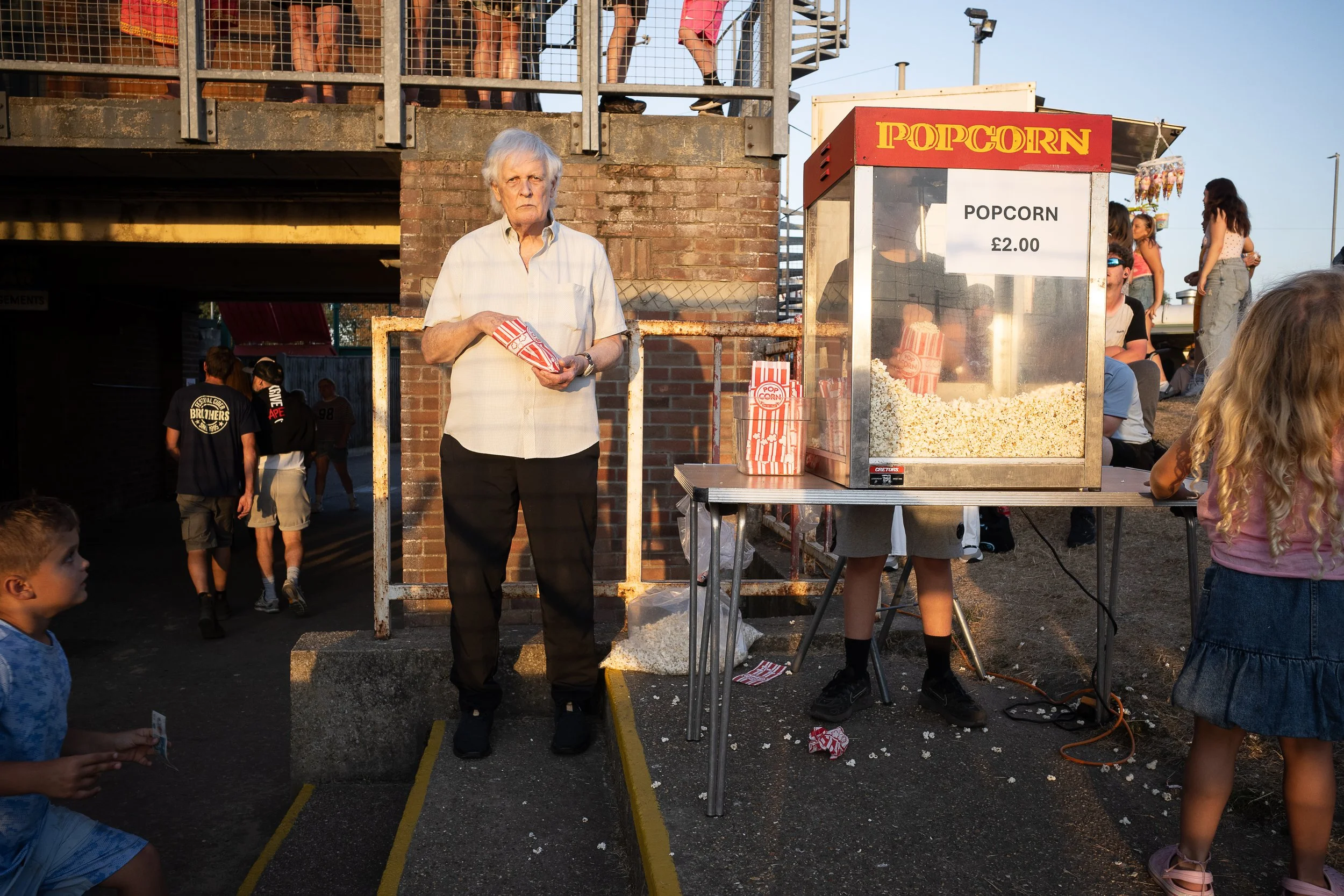 An elderly man standing at a popcorn stand holding a bag of popcorn at an outdoor fair or festival. Several people are around, with some waiting in line and others sitting and chatting.