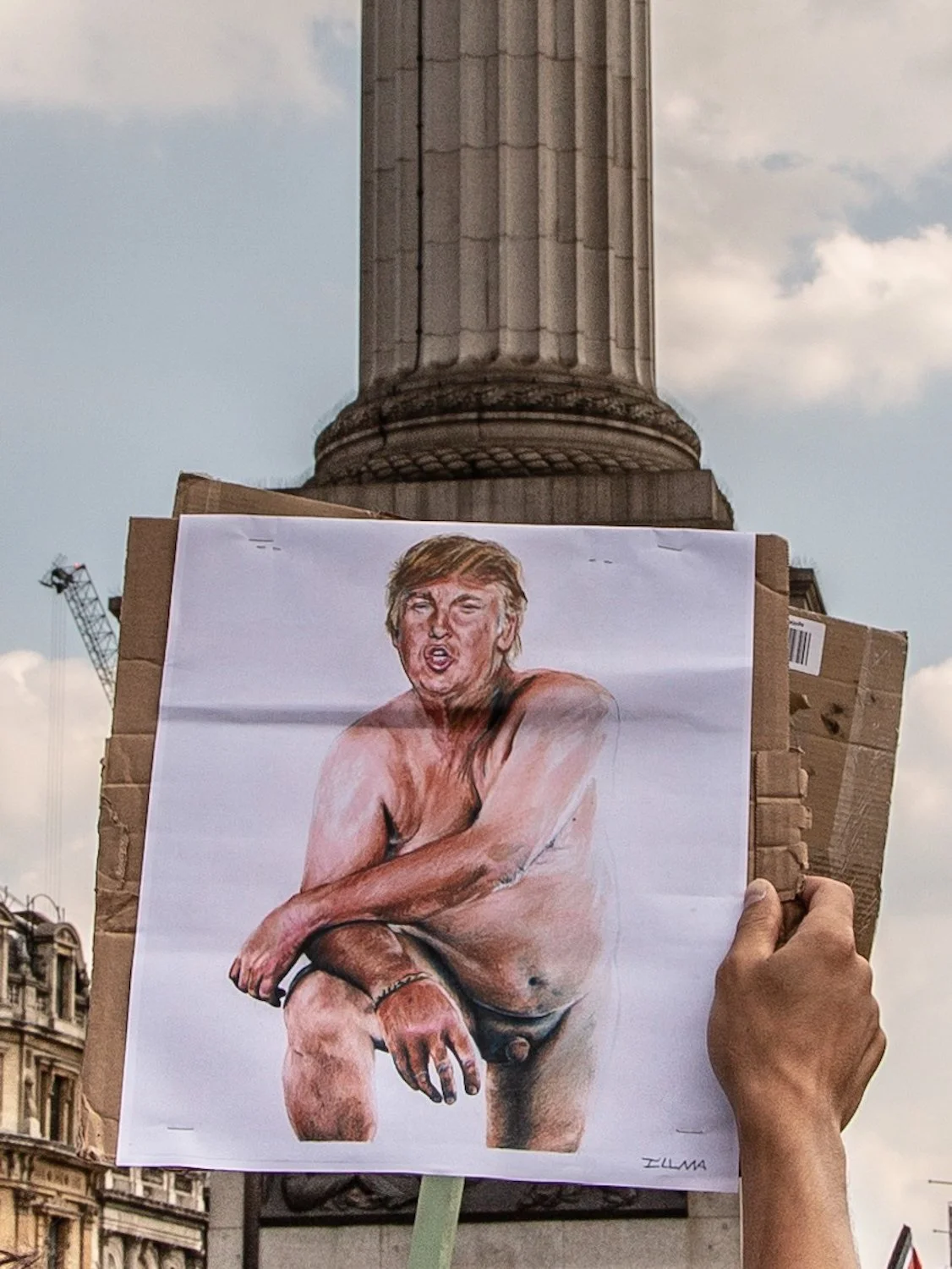 A hand holding a sign with a caricature of Donald Trump squatting, with a background of buildings and a partly cloudy sky.