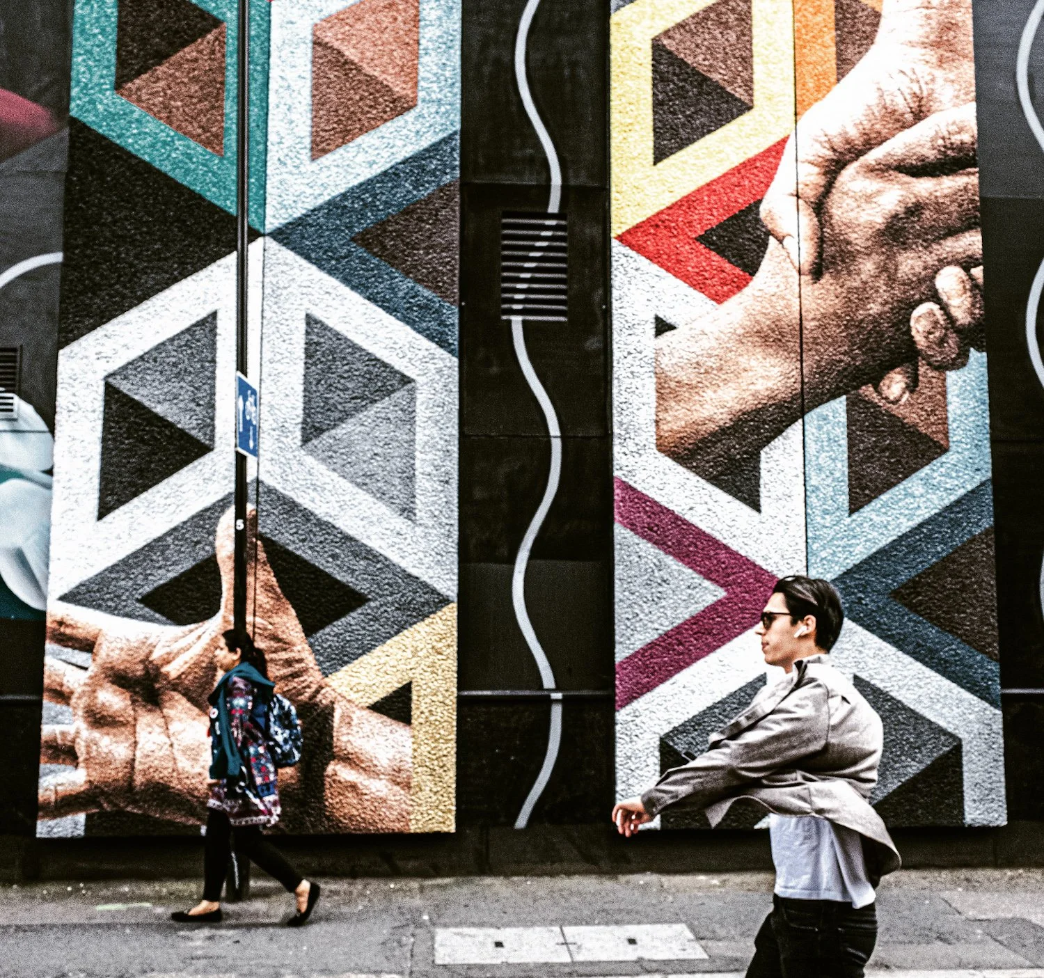 Urban street scene with a large colorful geometric mural on a building wall, featuring squares and triangles, and a mural of a person's face on the right. Two people are walking and one is sitting on the sidewalk.