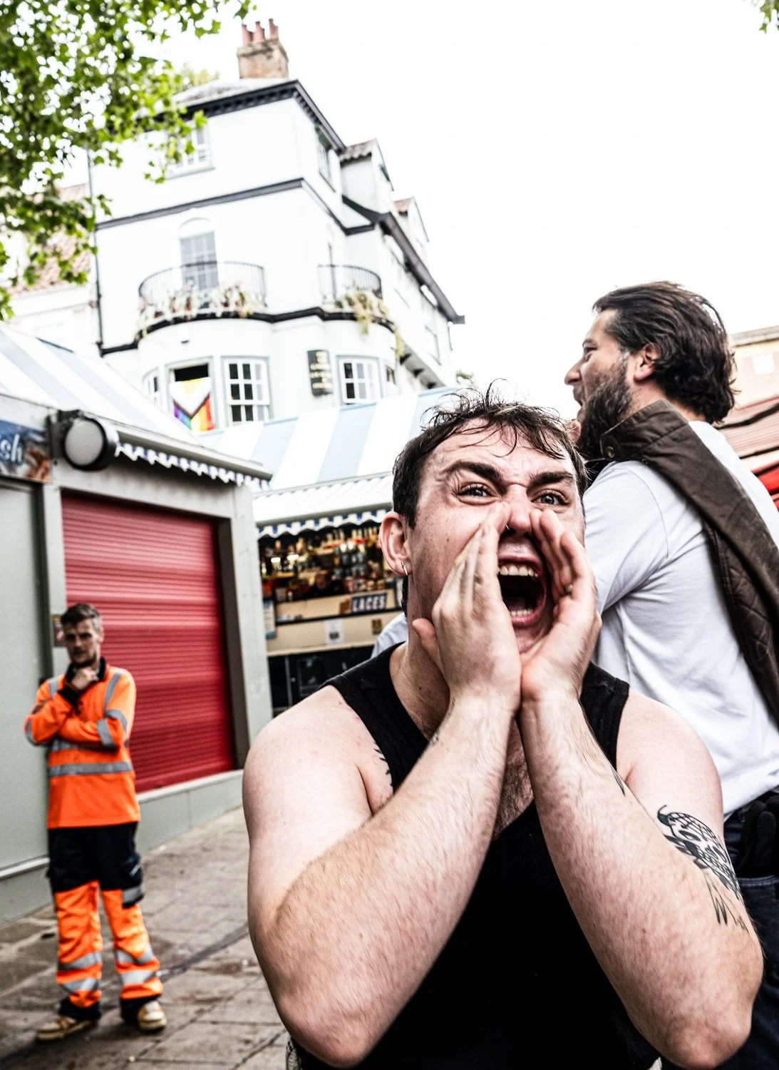 A person shouting with hands around their mouth on a street, with two other people, one in an orange safety vest, and a person with a beard and dark hair in the background. There are buildings and a market stall in the background.