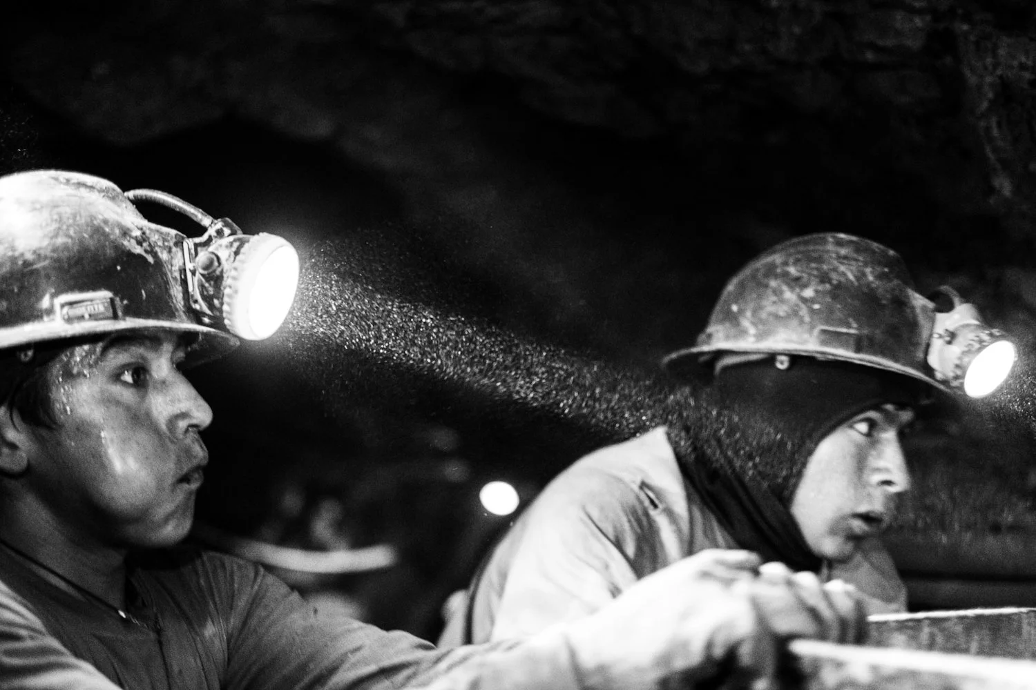 Two miners wearing helmets with headlamps working inside a dark mine shaft.