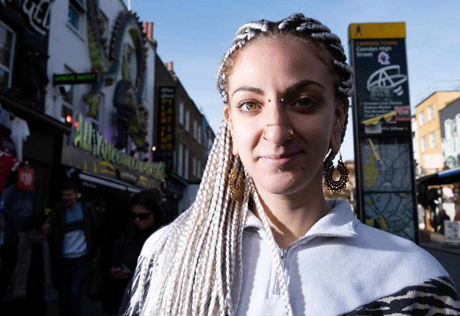 A woman with white braided hair, multiple facial piercings, and large earrings standing outdoors in a city street with shops and people in the background.