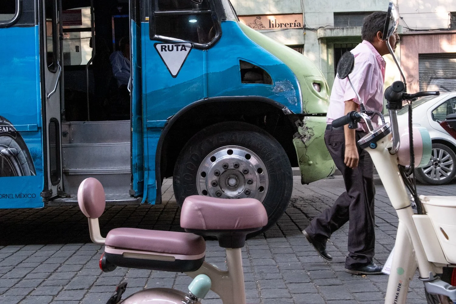 A man walking past a damaged bus with a light green front and blue body, parked on a city street, with a small pink and white scooter and a pink bicycle in the foreground.