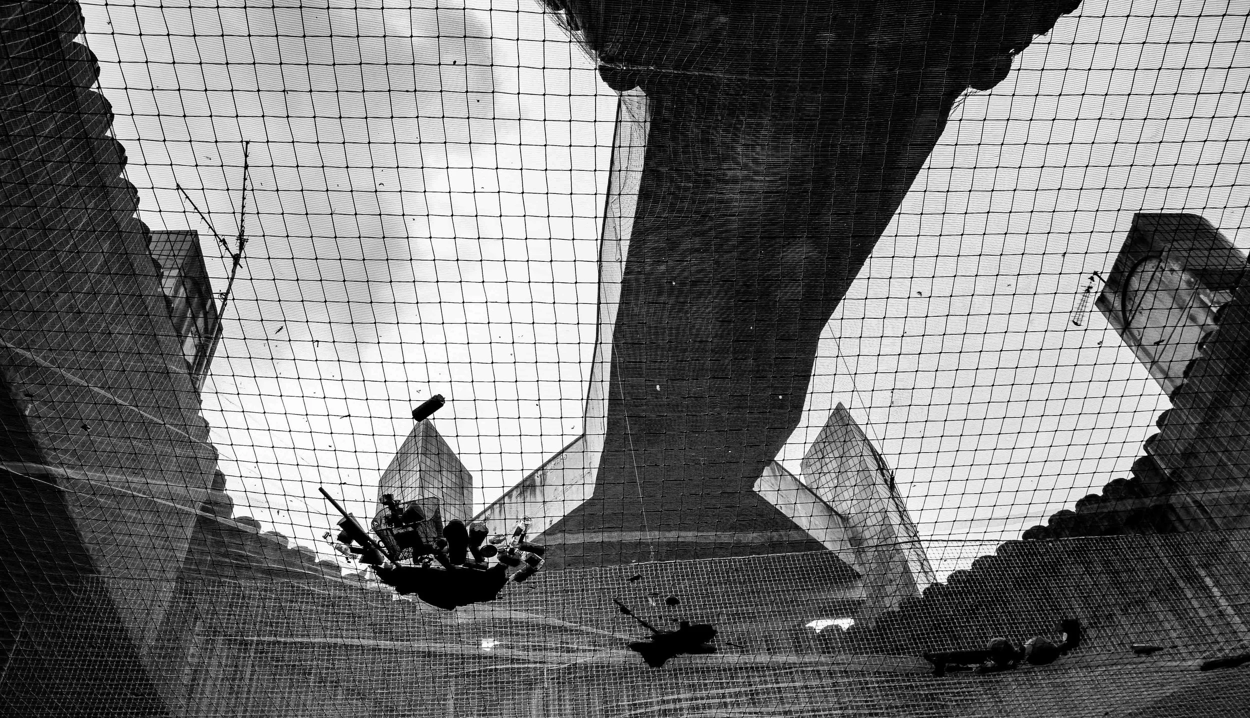 A black and white photo taken from below looking up at a cityscape framed by a netted canopy. Tall buildings and an overcast sky are visible.