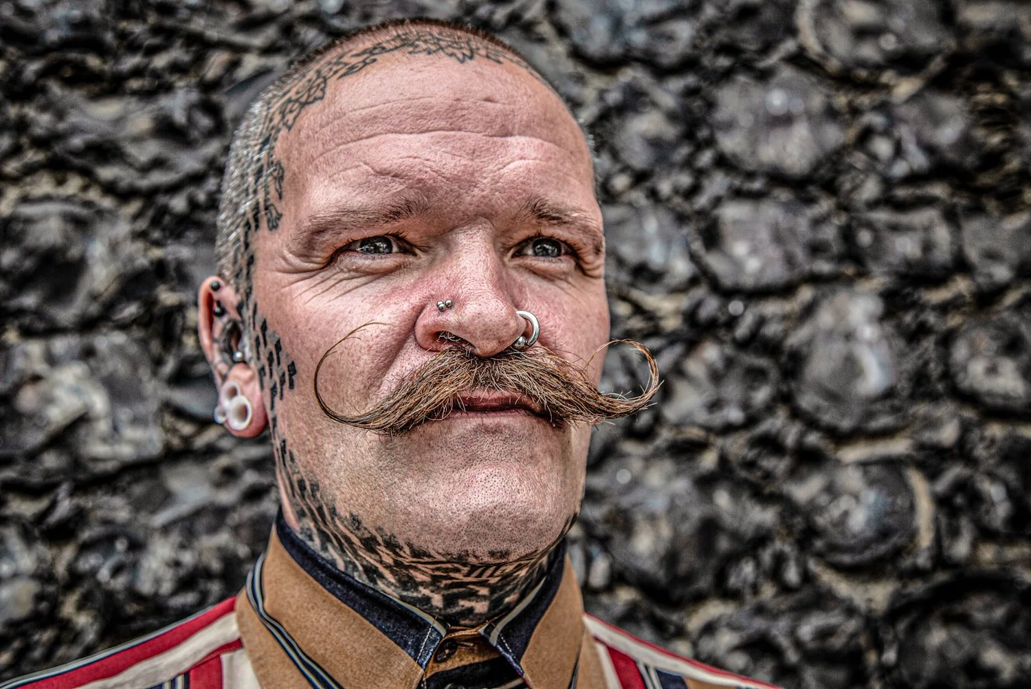 Close-up of a man with tattoos and multiple piercings, including nose and ears, with a styled mustache, standing outdoors on rocky terrain.