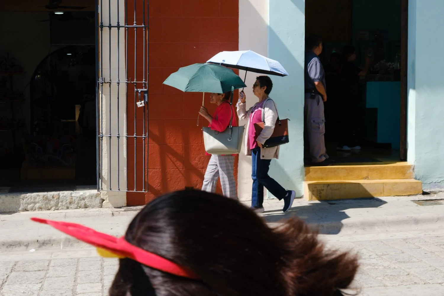Two women walking on the sidewalk in front of a building, each holding umbrellas, one pink and one white, under bright sunlight. The woman on the left is wearing a red jacket and checkered pants, while the woman on the right is dressed in a beige coa