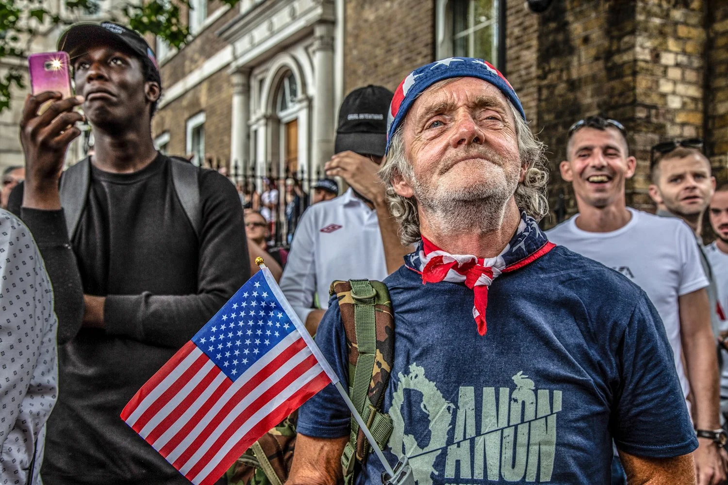 An older man with gray curly hair and a beard, wearing a bandana with the American flag pattern and a blue T-shirt with a military-themed design, holding a small American flag. He is surrounded by a group of young men, some of whom are smiling, in fr