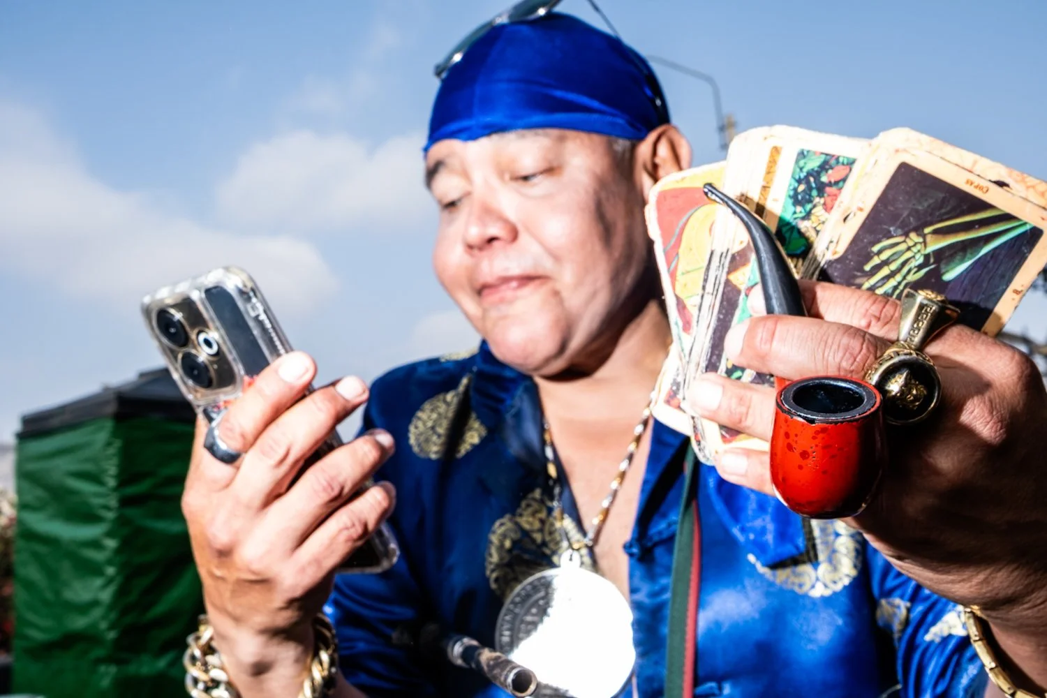 A person wearing a blue headscarf and jacket is holding a smartphone and a fan of tarot cards while looking at the phone.