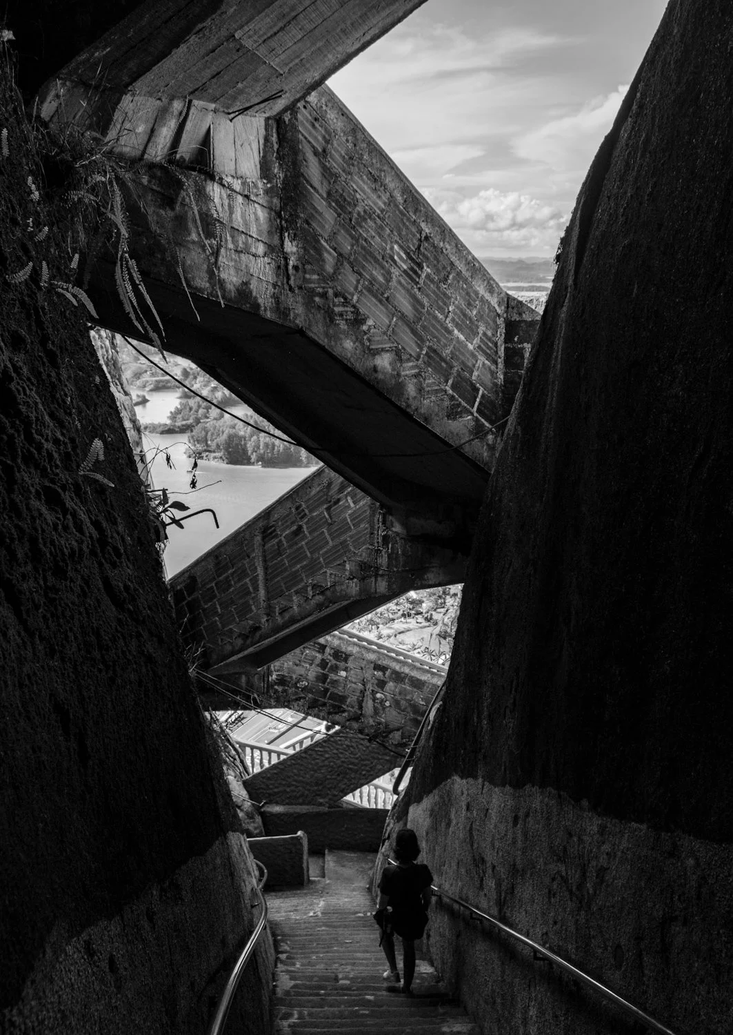 A person walking down a narrow outdoor stairway between large rock formations with large concrete bridges overhead, overlooking a landscape with water and trees in the distance.