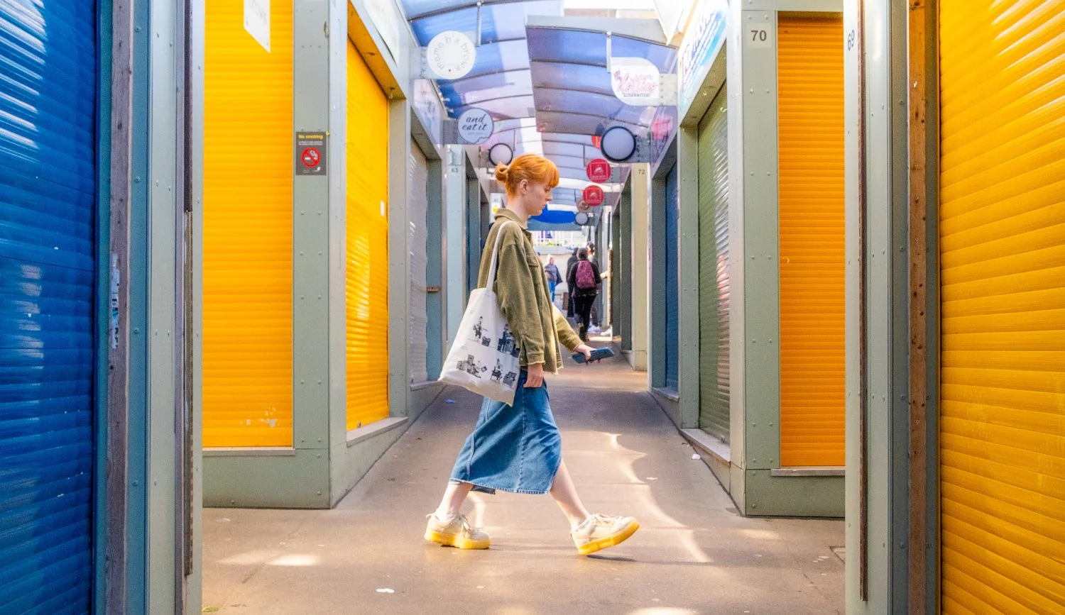 A young woman with red hair walking through a colorful outdoor shopping arcade with yellow, blue, and green closed shutters.