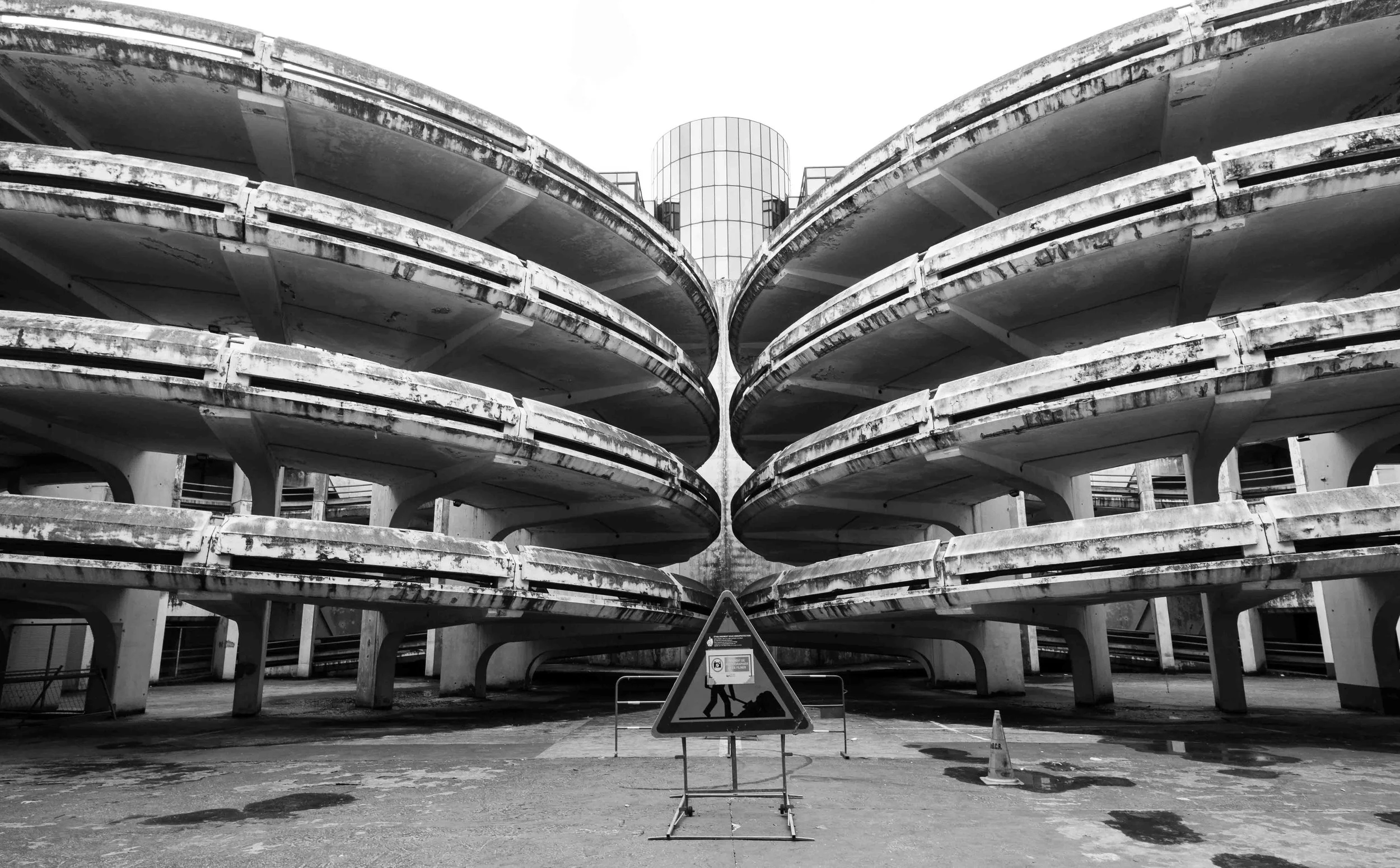 Black and white photo of a multi-level parking garage with concrete structure and a building with a rounded glass facade in the background. There are warning signs and a traffic cone in the foreground.