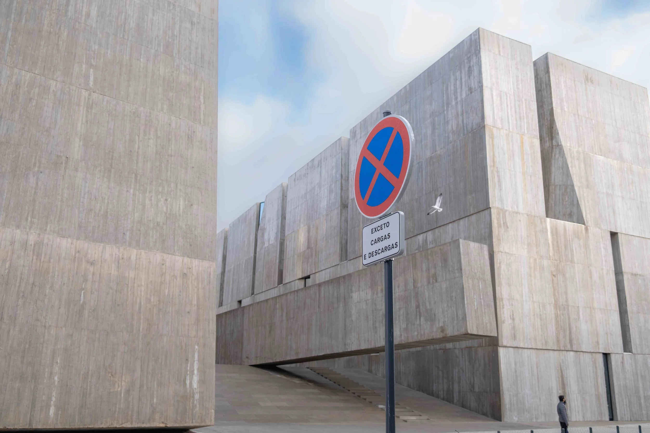 Modern concrete building with large geometric blocks and a no parking sign in front. The sky is partly cloudy.