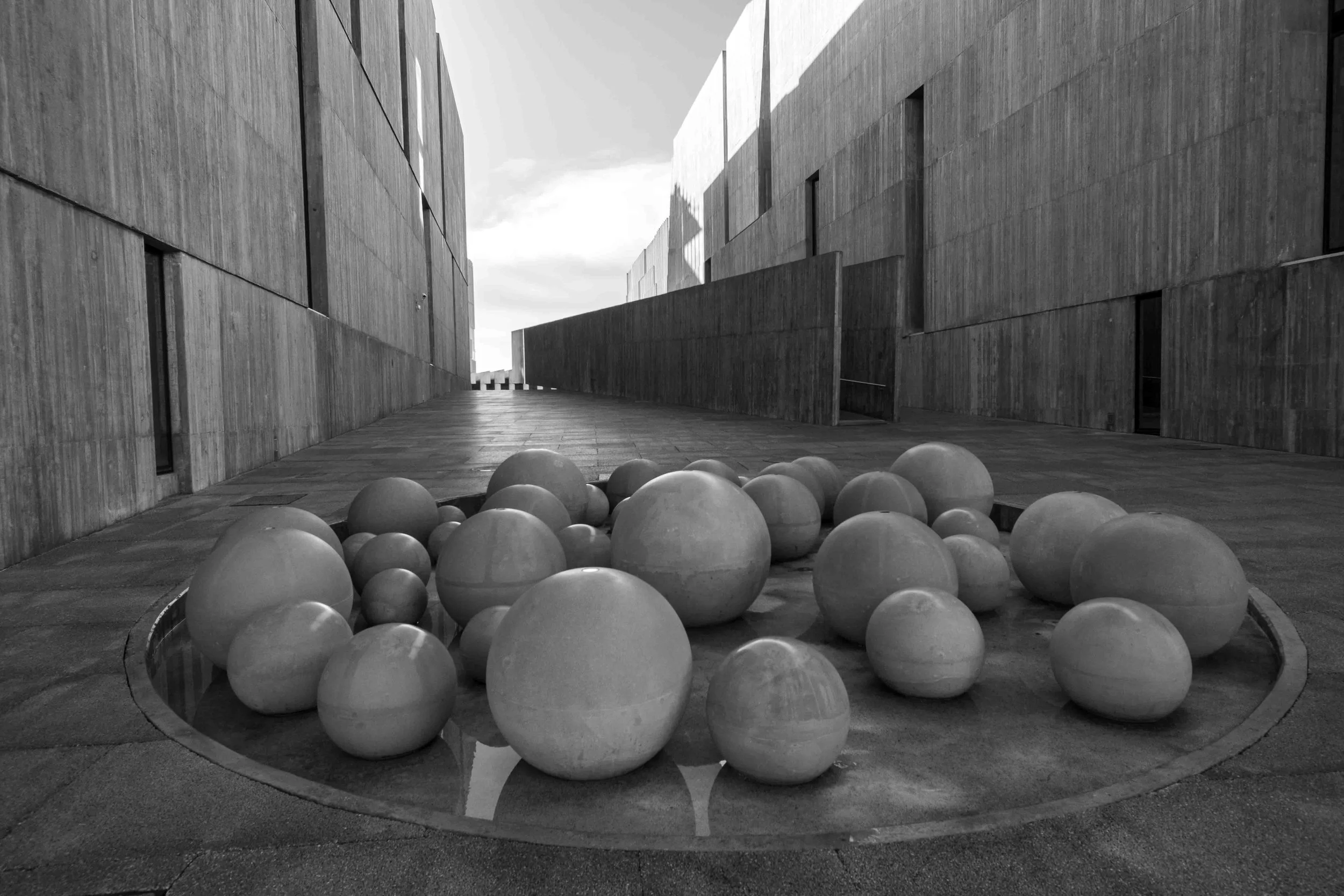 Black and white photo of a modern architectural building with a pathway in between. There are several spherical objects surrounding a small circular reflection pool in the foreground.