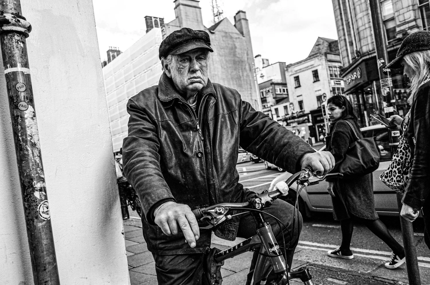 A black-and-white photo of an older man with a stern expression, wearing a leather jacket and a cap, standing next to a bicycle on a city street, with pedestrians and buildings in the background.