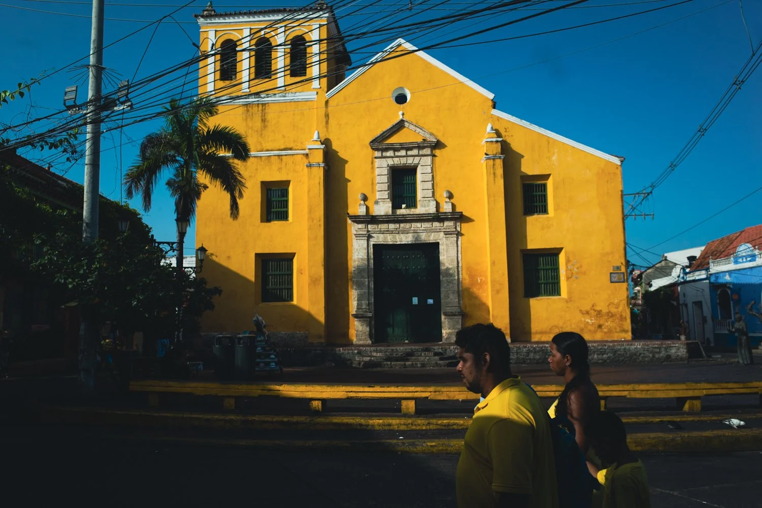 A yellow church with a baroque-style facade, dark green windows, and a bell tower, located in a sunny street with a palm tree and people walking by.