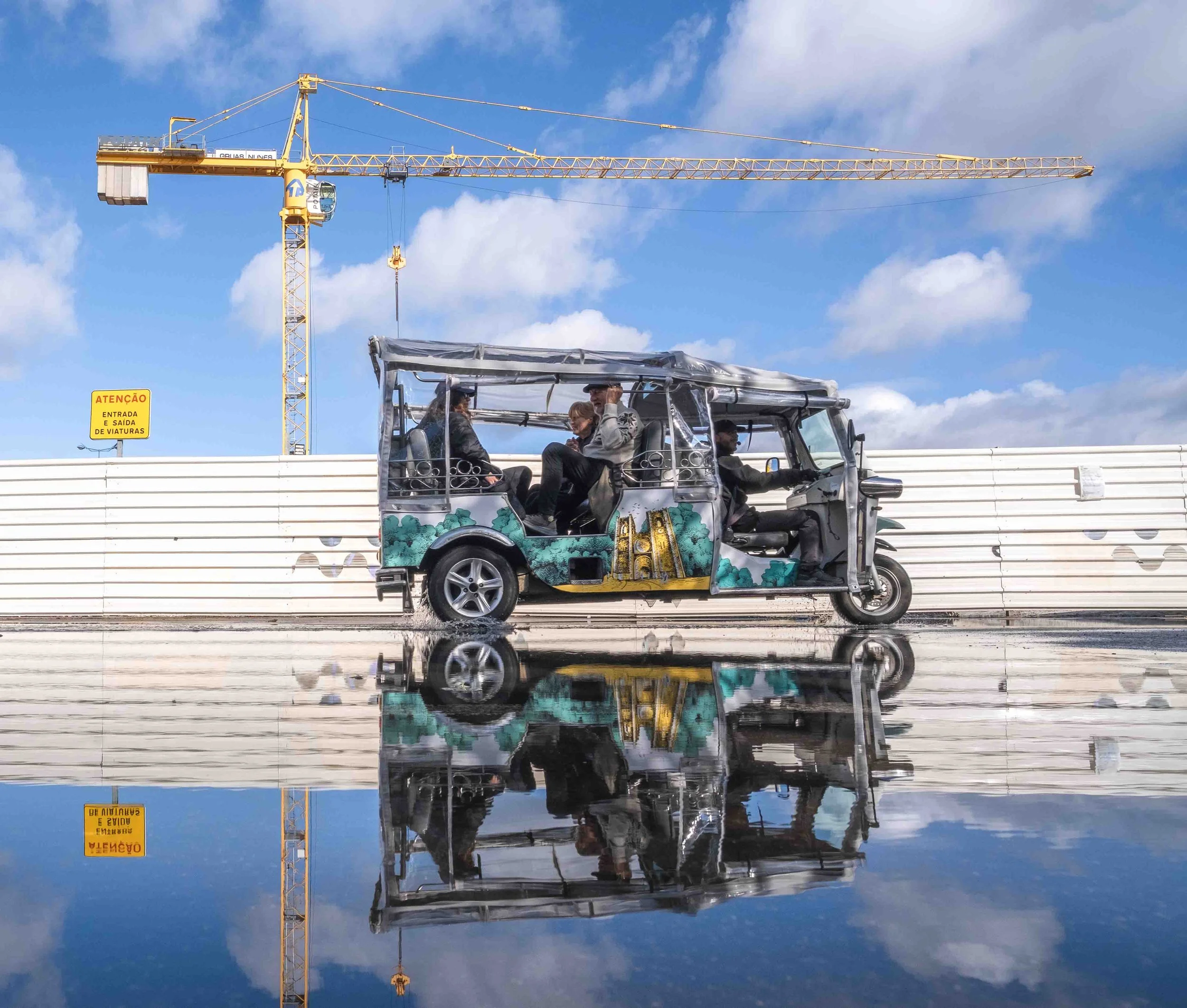 Yellow construction crane against blue sky with blue and white clouds, reflecting in a puddle, with a small vehicle carrying people in the foreground.