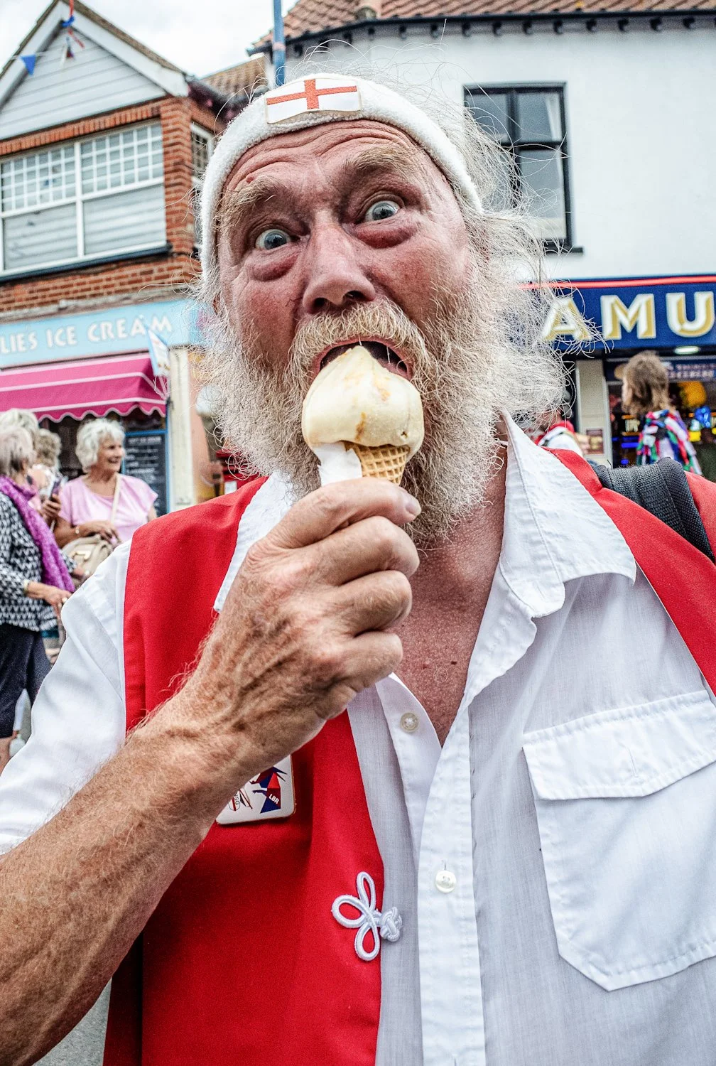 An elderly man with a white beard and a Suffolk headband is eating ice cream on a cone, standing outdoors in a busy street with shops and people in the background.