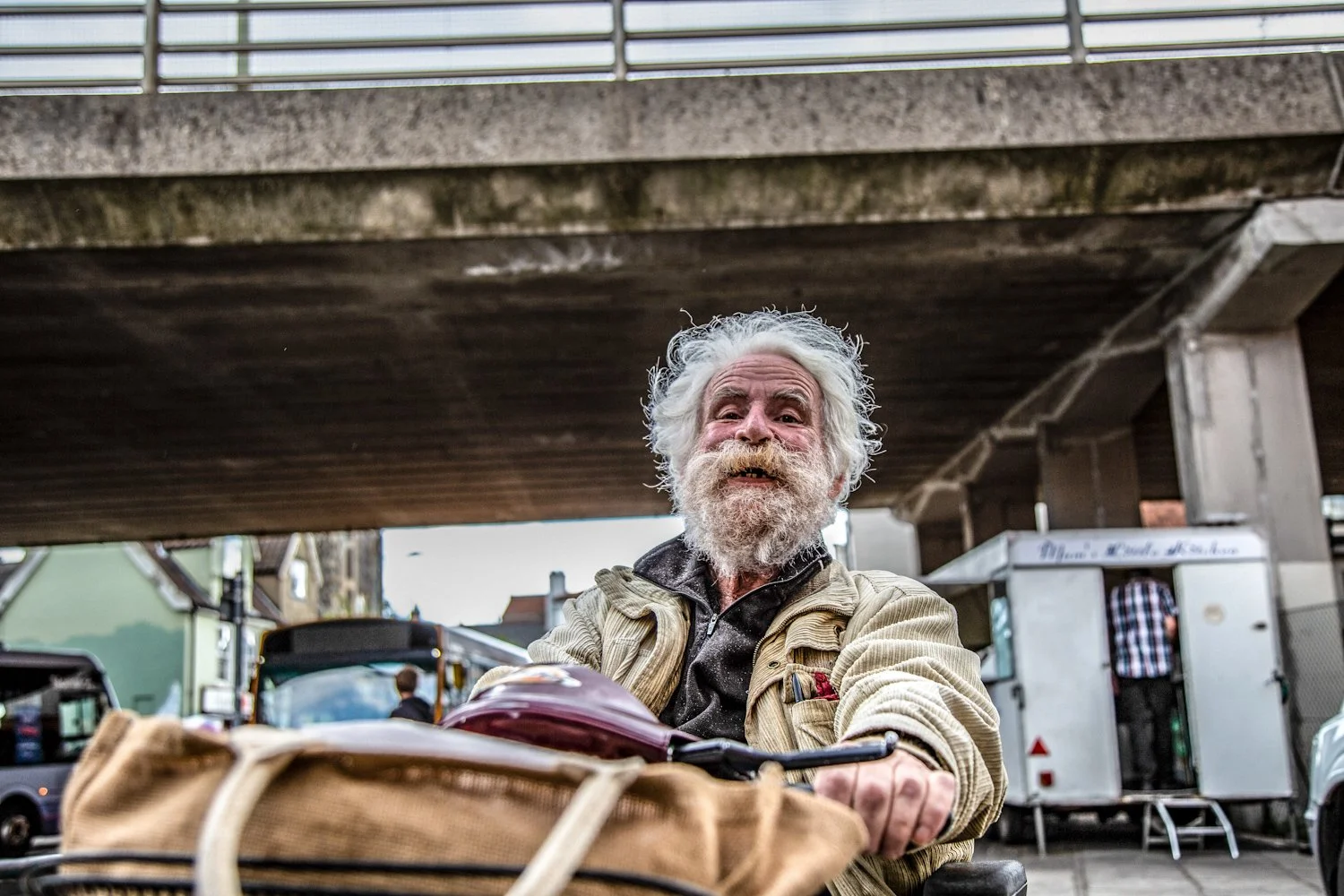 An elderly man with white hair and a beard smiling while sitting behind a bicycle in an urban setting under a bridge.