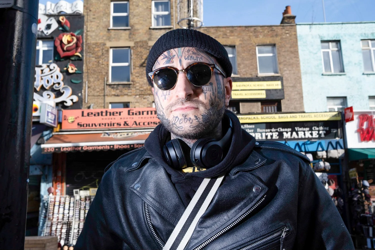 A man with tattoos on his face and neck, wearing a black beanie, sunglasses, a black leather jacket, and headphones around his neck, stands outdoors in front of shops and buildings.