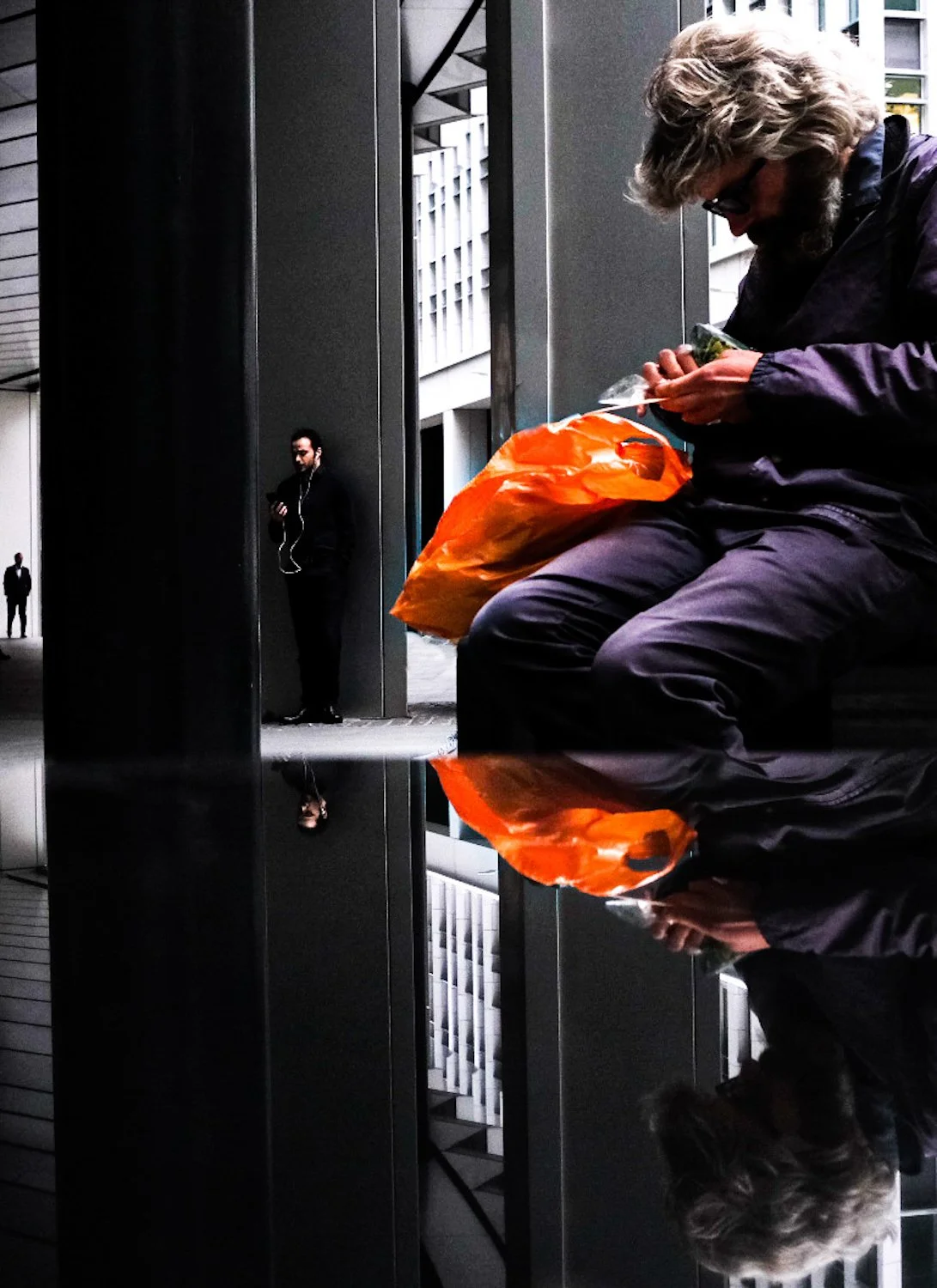 A man with gray hair and sunglasses sitting on a bench, looking at his phone, with an orange shopping bag beside him. In the background, a man with a beard dressed in black, holding a phone and listening to earbuds, stands near a dark wall.