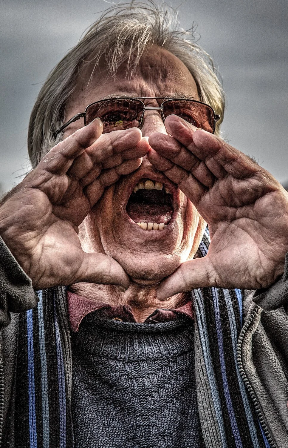 An elderly man with gray hair, wearing sunglasses and a striped sweater with a zip-up jacket, shouts or calls out loudly with his mouth open, forming a circle with his hands around his mouth.