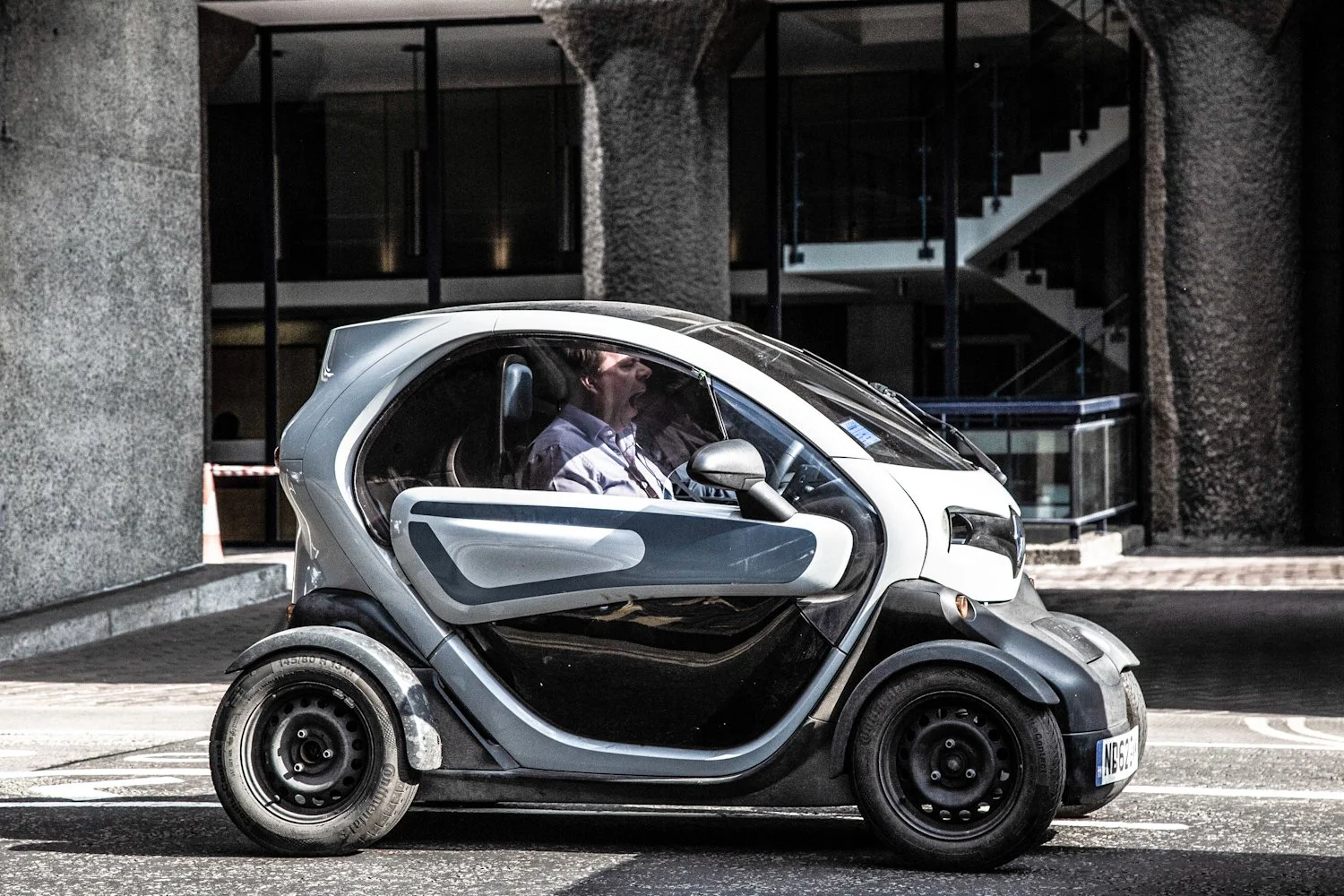 A small, modern, electric car with a transparent bubble-shaped cabin, driven by a person yawning inside, on a city street with contemporary buildings in the background.