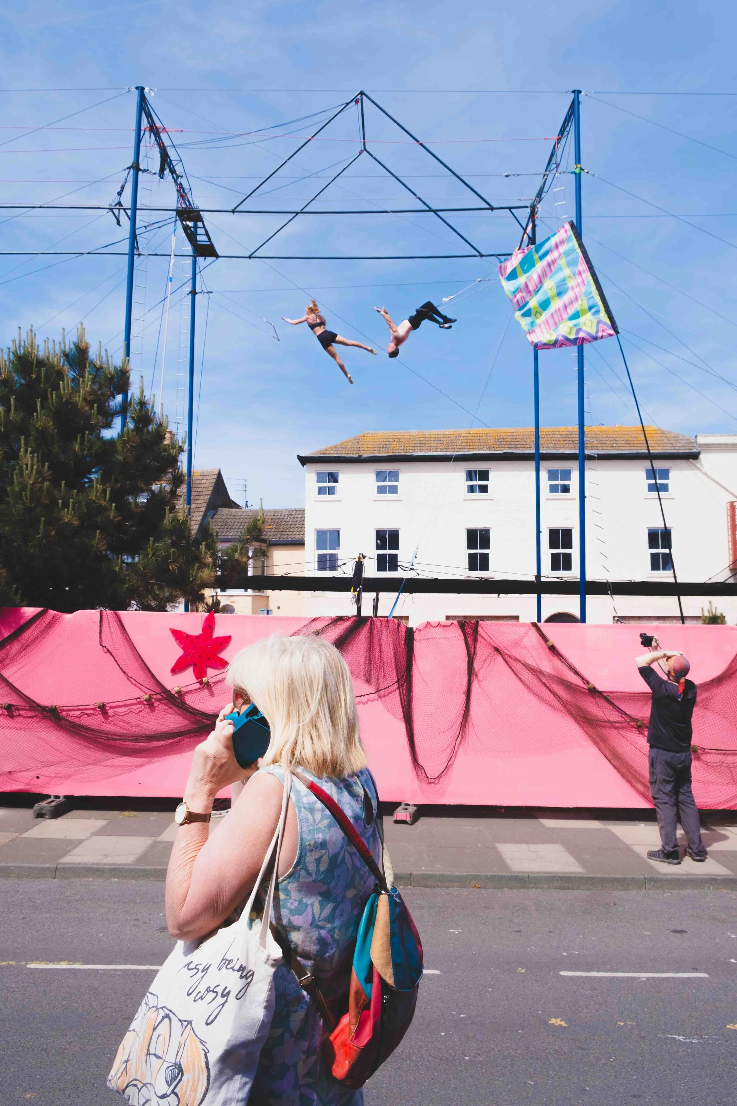 People watching as two aerial performers perform on a trapeze structure in an outdoor street scene. In the foreground, a woman with white hair is talking on a cell phone, and a man is taking photos of the performance. The background includes buildings, a tree, and a pink decorative barrier.