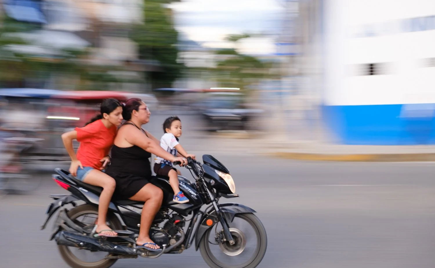 Three people riding a black motorcycle on a city street, with a woman in black in the middle, a girl in red on the back, and a boy in white in front, all wearing casual clothes.