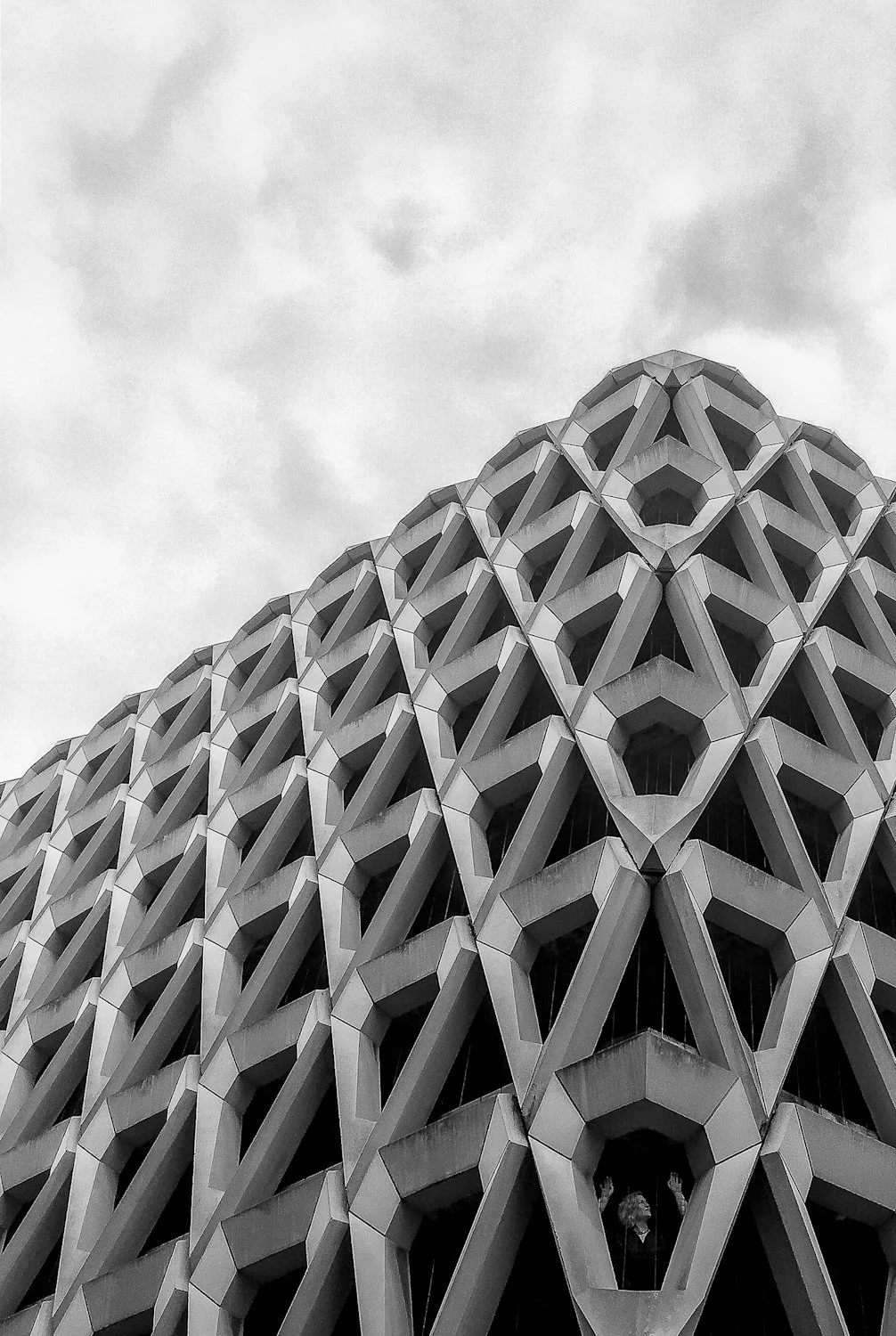 Low-angle black and white photo of a modern building with a geometric honeycomb-like facade and a person looking out from one of the windows.