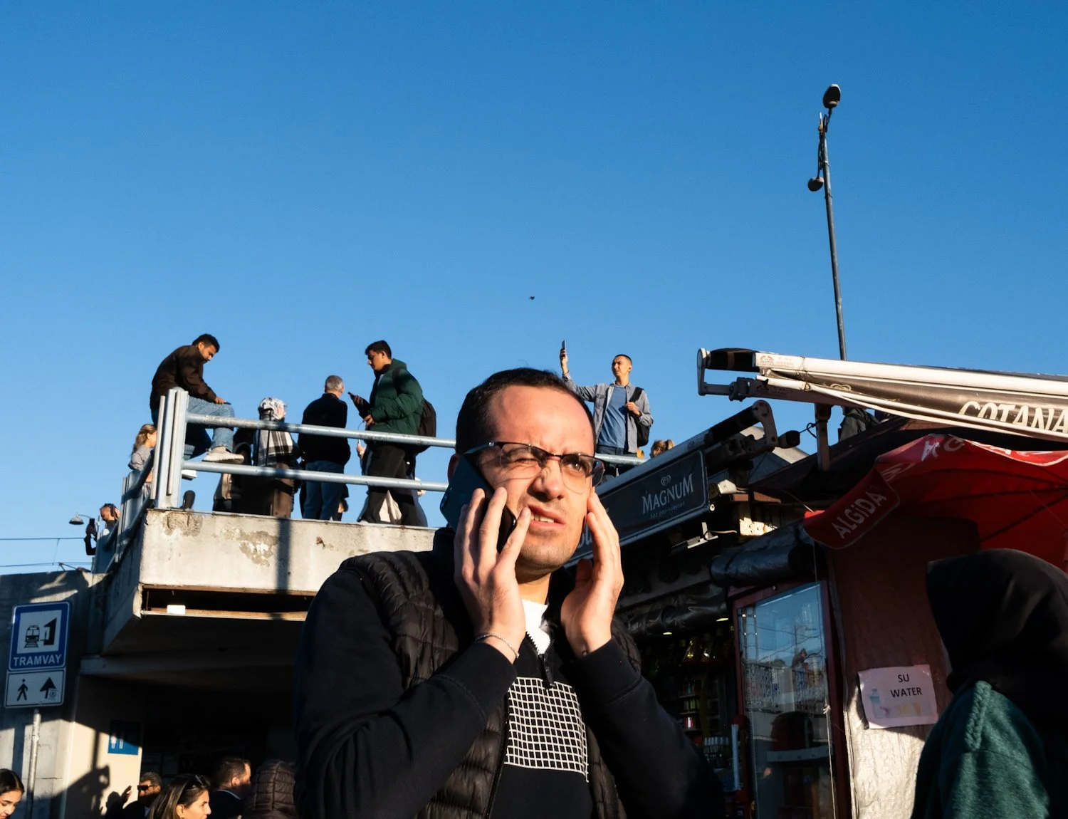 A man with glasses and dark hair holding a cell phone to his ear, with a confused or distressed expression, standing in a busy outdoor area with a group of people standing on an overpass or balcony above him.