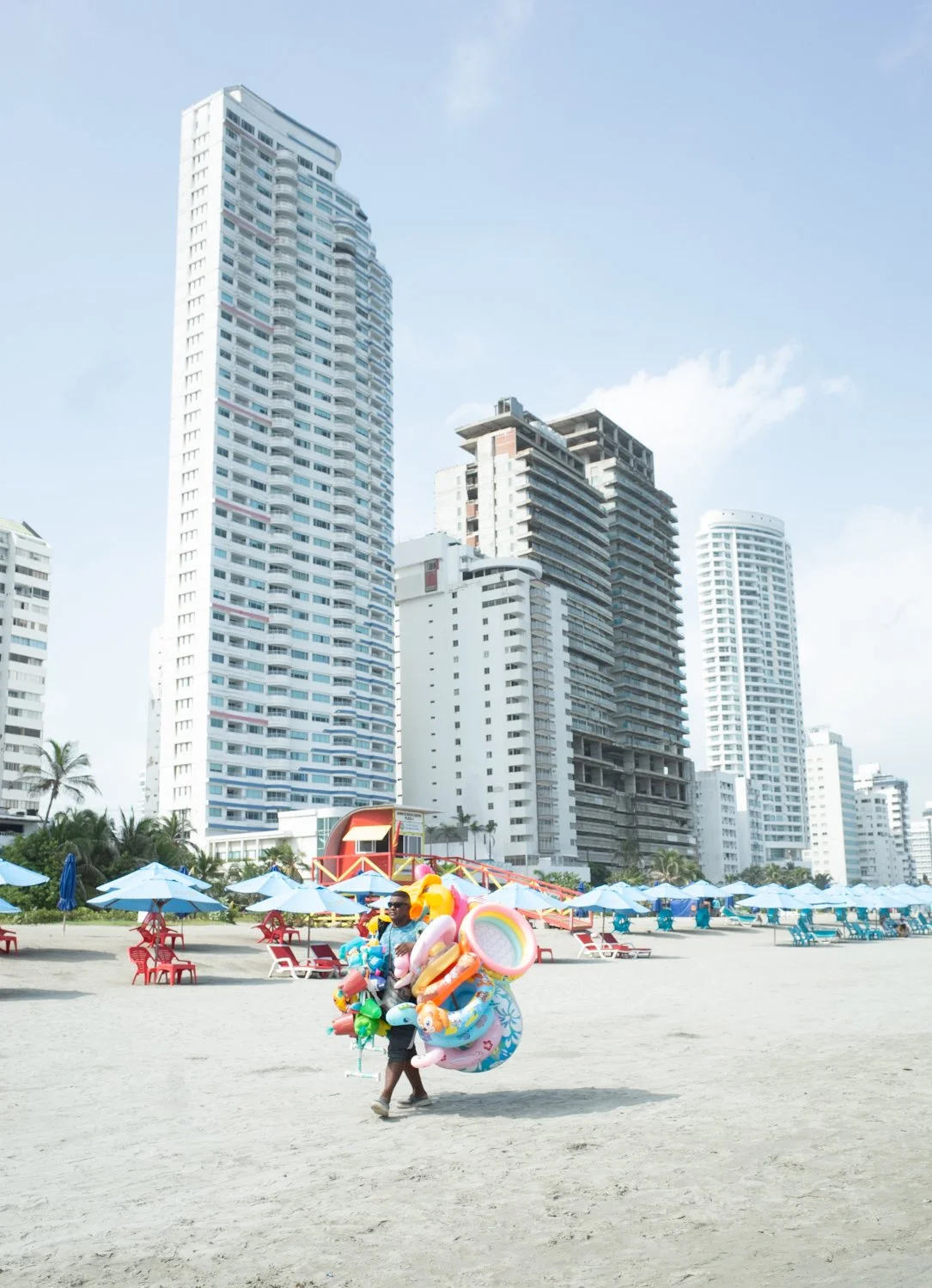 A man carrying pool floaties and toys on a sandy beach. Behind him, there are beach chairs with umbrellas and tall buildings in the city skyline.